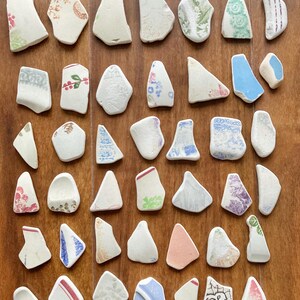 May include: A collection of ceramic fragments with diverse patterns and colours, including floral and striped designs. The pieces are in shades of white, blue, pink, and green, arranged on a wooden surface.