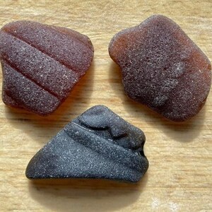 May include: Three pieces of sea glass, one dark grey, two brown. The brown pieces have a striated texture.