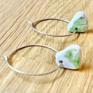 May include: A pair of hoop earrings with white and green marbled ceramic pendants. The earrings have silver-colored hoops and are displayed on a wooden surface. The pendants have a triangular shape.