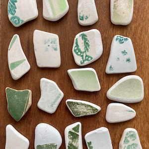 Pode incluir: Uma coleção de 18 azulejos de cerâmica branca com desenhos pintados em verde. Os azulejos são de várias formas e tamanhos e têm um aspeto rústico e feito à mão.