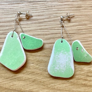 May include: A pair of handmade earrings featuring a light green, irregular shape with a white border. The earrings are connected by a silver wire and post. The earrings have a unique, artistic design.
