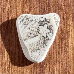 May include: A white, heart-shaped piece of sea pottery with a black floral pattern. The pottery has a smooth, worn texture and is set against a brown wooden surface. The design includes flowers and other decorative elements.