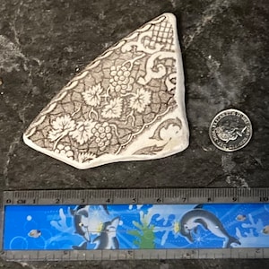 May include: A white ceramic shard with a brown floral design featuring grapes and leaves. The shard is approximately 12.5 cm long and 7.5 cm wide.