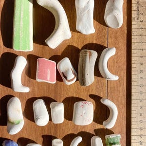Könnte beinhalten: Eine Sammlung kleiner, unregelmäßig geformter, weißer und farbiger Objekte, möglicherweise Strandsteine oder Kunstwerke, auf einer Holzoberfläche angeordnet. Einige Teile haben grüne, rosa oder blaue Akzente. Ein Lineal und zwei Münzen sind ebenfalls vorhanden.