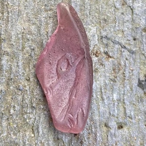 May include: A piece of sea glass in a soft, translucent pink hue. The glass has a smooth, rounded shape with a textured surface and appears to have an etched design. The sea glass is resting on a rough, grey stone surface.
