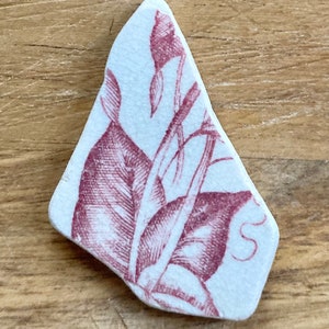 May include: A white ceramic tile with a red floral design. The design features a stylized flower with leaves and a stem. The tile has a slightly irregular shape.