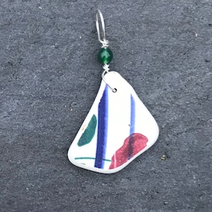May include: A white ceramic earring with blue and green painted designs. The earring is attached to a silver wire with a green bead.