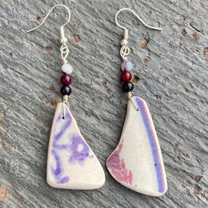 Peut inclure: Boucles d'oreilles faites à la main. Chaque boucle d'oreille est ornée d'un crochet argenté et d'une chaîne de perles colorées. Les pendentifs en céramique blanche présentent des motifs violets et roses. Un accessoire artistique unique.