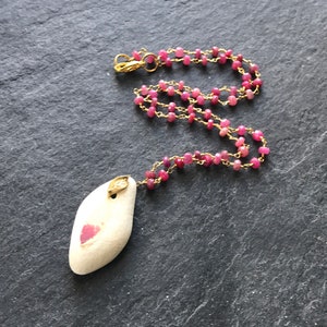以下が含まれることがあります： ピンクのハートのデザインが施された白い楕円形のペンダントが、ピンクのビーズが付いたゴールドチェーンからぶら下がっています。チェーンにはゴールドの留め具が付いています。