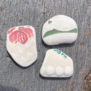 May include: Three decorative beach stones with painted designs. One stone features a red floral pattern, another has a green stripe, and the third has three turquoise circles. The stones are white and have a smooth texture.