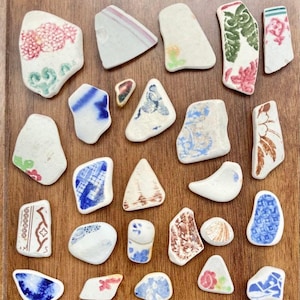 May include: A collection of sea pottery shards with various patterns and colors. The pieces feature floral, geometric, and abstract designs in shades of blue, red, green, and brown. The pottery fragments are arranged on a wooden surface.