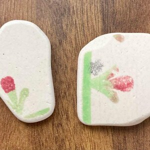 May include: Two off-white ceramic stones with floral designs. One stone features a red flower with green leaves, the other has a similar flower with a green stem and additional floral elements. The stones are on a wooden surface.