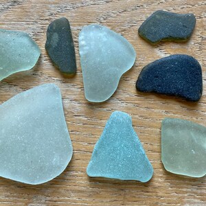 Puede incluir: Una colección de ocho piezas de vidrio de mar en varios tonos de verde y azul, junto con dos piedras gris oscuro. Las piezas de vidrio de mar son lisas y tienen un aspecto esmerilado.