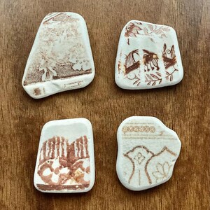Peut inclure: Quatre morceaux de poterie ancienne avec des motifs floraux et paysagers bruns sur fond blanc. Les éclats de poterie ont des formes irrégulières et sont disposés sur une surface en bois.