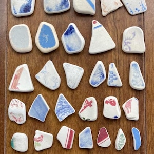 Può includere: Una collezione di trentotto frammenti di vetro marino e ceramica di varie forme e dimensioni. I pezzi presentano basi bianche con motivi decorativi in blu, rosso, verde e marrone, caratterizzati da bordi levigati e arrotondati.