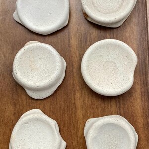 Op de afbeelding: Zes kleine, ronde, crèmekleurige keramische schaaltjes met onregelmatige randen zijn gerangschikt op een houten oppervlak. De schaaltjes hebben een getextureerde uitstraling en een ondiep, concaaf ontwerp, wat suggereert dat ze voor kleine porties zijn.