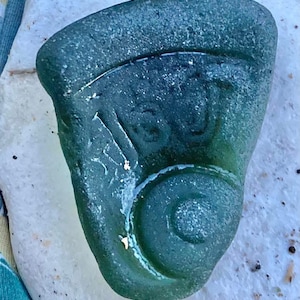 May include: A piece of sea glass in a teal color, with a design etched into the surface. The design includes a spiral and other curved lines. The glass has a textured appearance and is set against a white background.