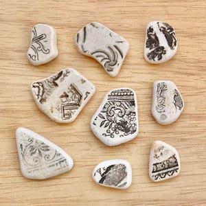 May include: Nine pieces of white ceramic with black floral and geometric patterns. The shapes vary, with some resembling pebbles. The pieces are arranged on a light brown wooden surface, showcasing the detailed designs.