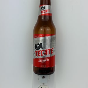 May include: A brown glass bottle of Tecate Original beer with a silver label and red accents. The bottle is plugged into a standard electrical outlet.