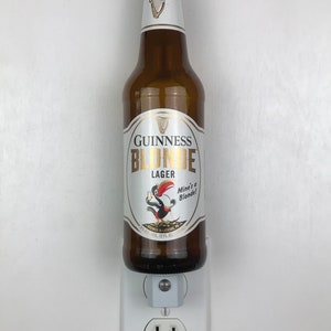 May include: A brown glass bottle of Guinness Blonde Lager with a white label featuring a cartoon toucan holding a beer. The label reads "Guinness Blonde Lager" and "Mine's a Blonde!" The bottle is plugged into a white electrical outlet.