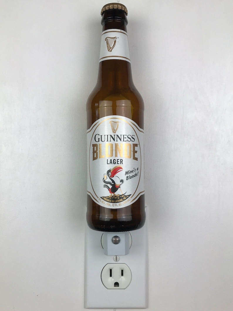 May include: A brown glass bottle of Guinness Blonde Lager with a white label featuring a cartoon toucan holding a beer. The label reads "Guinness Blonde Lager" and "Mine's a Blonde!" The bottle is plugged into a white electrical outlet.
