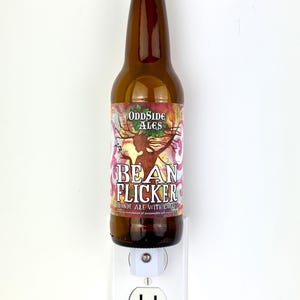 May include: A brown glass beer bottle with a silver cap is plugged into a white electrical outlet. The bottle has a colorful label that reads "BEAN FLICKER" and "Blonde Ale with Coffee". The product is a night light.