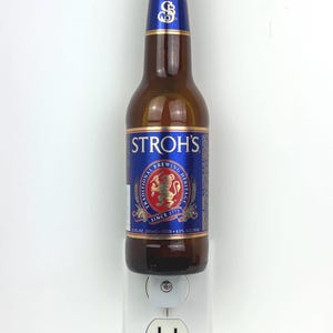 May include: A brown glass bottle of Stroh's beer with a blue label. The label features the Stroh's logo, a lion, and the text "Traditional Brewing Heritage Since 1775". The bottle is plugged into a white electrical outlet.