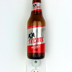 May include: A brown glass Tecate beer bottle with a red and silver label featuring an eagle logo and the text Original 12 FL. OZ. The bottle is mounted onto a white electrical wall outlet plate with a single circular plug-in port at the top.
