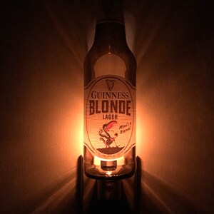 May include: A glass bottle night light shaped like a Guinness Blonde Lager beer bottle. The bottle has a white label with the text "Guinness Blonde Lager" and a cartoon bird with the text "Mine's a Blonde!"