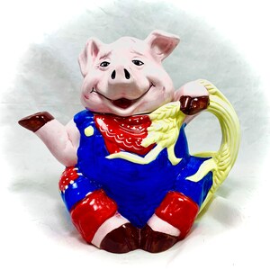 Könnte beinhalten: Eine Keramik-Teekanne in Form eines Schweins, das eine Latzhose und ein rotes Bandana trägt. Die Teekanne hat einen Henkel aus gelbem Weizen.