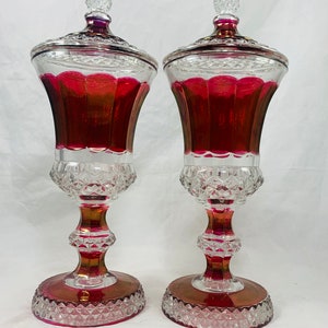 Może przedstawiać: Para vintage'owych cukierniczek z rubinowego szkła z przezroczystymi szklanymi pokrywkami i podstawami ze szkła ciętego w kształcie diamentu.