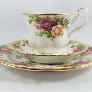 May include: A white teacup and saucer set with a floral pattern of pink and yellow roses. The set has a gold trim around the edges.