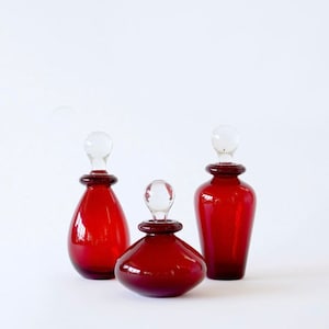 May include: Three glass bottles with clear glass stoppers. The bottles are a deep red colour and are of varying sizes.