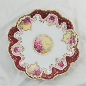 May include: A white porcelain plate with a burgundy rim and gold accents. The plate features a floral design with pink and white roses.