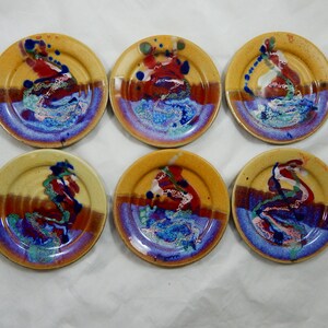 May include: Set of six small, round ceramic plates. Each plate features a unique abstract design with a blend of colors including yellow, red, blue, and brown. The plates have a glossy finish and are suitable for serving small portions.