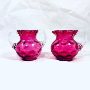 Puede incluir: Dos jarras de cristal color arándano con asas transparentes en forma de bucle. Las jarras tienen un borde con volantes y un patrón de círculos en relieve. El cristal es de un color rosa intenso y las asas son transparentes.