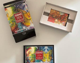 Op de afbeelding: Het Osho Zen Tarot-deck bevat een verticale doos met kleurrijke illustraties, een horizontaal instructieboek met bijpassende omslag en een witte kartonnen tray met een stapel kaarten voorzien van abstracte cirkelpatronen en zwarte lijntekeningen.