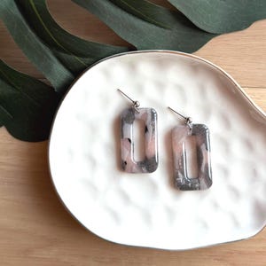 May include: A pair of rectangular earrings with a marbled design in shades of pink, gray, and white. The earrings are displayed on a white, organically shaped dish. The background includes a large green leaf and a wooden surface.