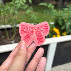 Könnte beinhalten: Ein rosa sticker in Schleifenform mit den Worten "Doing my best" in kursiver Schrift. Der Aufkleber hat rote vertikale Streifen und wird in der Hand einer Person vor einem verschwommenen Hintergrund aus Grün gehalten.