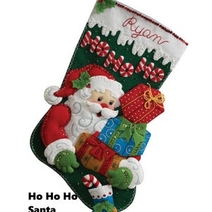 Pode incluir: Meia de Natal de feltro verde com uma aplicação de Papai Noel segurando presentes. A meia tem as palavras "Ho Ho Ho" e o nome "Ryan" bordado.