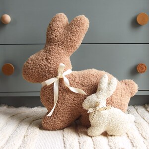May include: Two plush bunny rabbit toys. The larger bunny is brown and has a white ribbon around its neck. The smaller bunny is white and has a black and white polka dot ribbon around its neck. Both bunnies are sitting on a white rug.