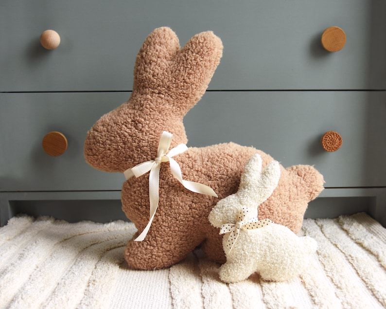 May include: Two plush bunny rabbit toys. The larger bunny is brown and has a white ribbon around its neck. The smaller bunny is white and has a black and white polka dot ribbon around its neck. Both bunnies are sitting on a white rug.