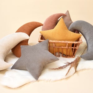 Op de afbeelding: Een set pluche kussens in de vorm van sterren en halve manen. De kussens zijn gemaakt van corduroy stof in verschillende kleuren, waaronder grijs, bruin, geel en roze. De kussens zijn gerangschikt op een wit nep bontkleed.