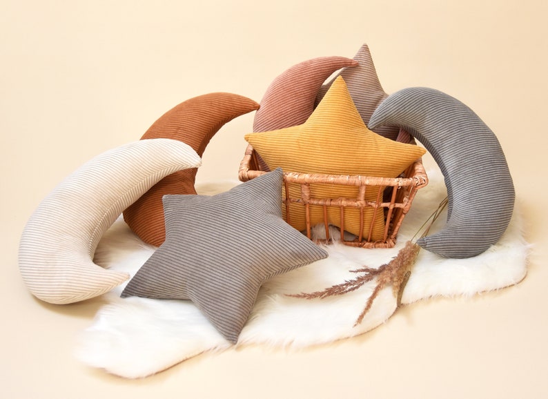 Op de afbeelding: Een set pluche kussens in de vorm van sterren en halve manen. De kussens zijn gemaakt van corduroy stof in verschillende kleuren, waaronder grijs, bruin, geel en roze. De kussens zijn gerangschikt op een wit nep bontkleed.