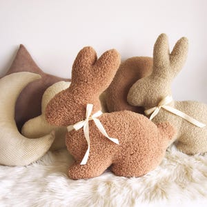 May include: Two plush brown bunny rabbit toys with cream colored ribbon bows, and a cream colored crescent moon shaped pillow with brown corduroy trim.