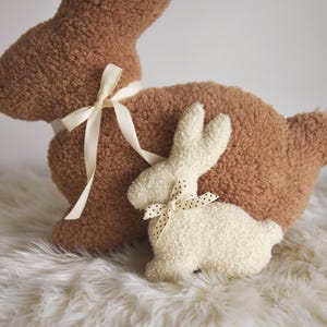 May include: Two plush bunny toys, one brown and one white. The brown bunny has a white ribbon tied around its neck. The white bunny has a black and white polka dot ribbon tied around its neck.