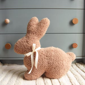 May include: A plush brown bunny toy with a white ribbon tied around its neck. The bunny is sitting on a white rug with a striped pattern.