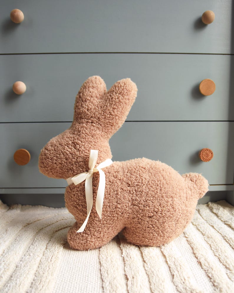 May include: A plush brown bunny toy with a white ribbon tied around its neck. The bunny is sitting on a white rug with a striped pattern.