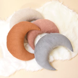 Moon shaped pillows rest at furry, white rug. They are made of corduroy fabric in bohemian colors: rust color, rose, beige and light grey.