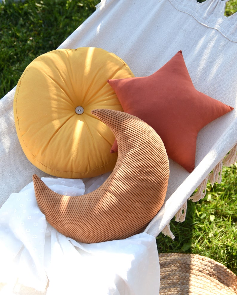 Op de afbeelding: Drie decoratieve kussens in een hangmat. Een kussen is rond en geel, een ander is een stervorm in een roodbruine kleur en de derde is een halve maanvorm in een bruine corduroy stof.